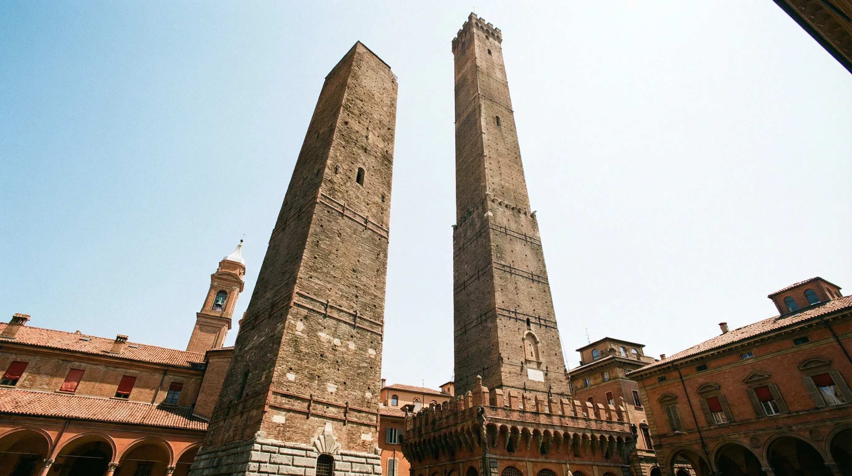 The tall medieval brick Asinelli and Garisenda towers rising above the red rooftops of Bologna under a bright sky.